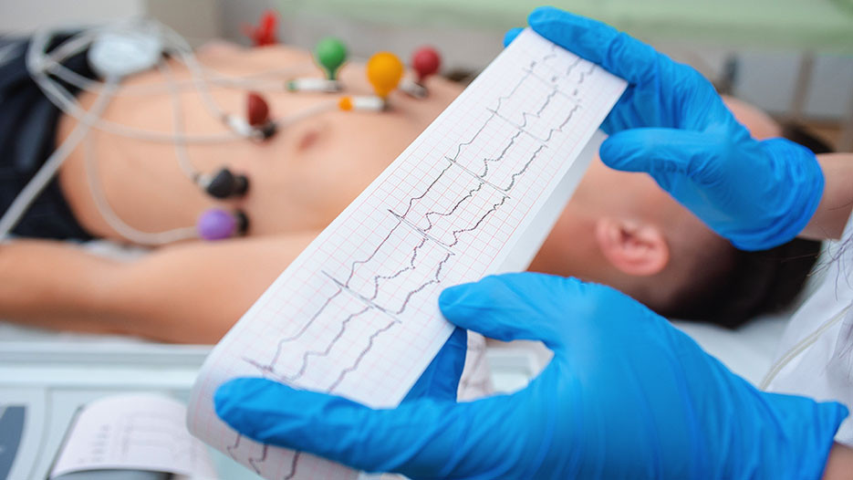 Gloved hands hold a printed ECG strip while a patient with chest electrodes lies in the background on a table.