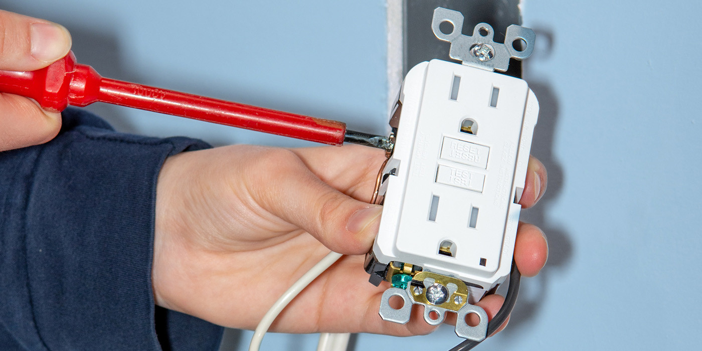 Hand uses a red screwdriver to secure wiring on a white GFCI electrical outlet mounted in a wall box