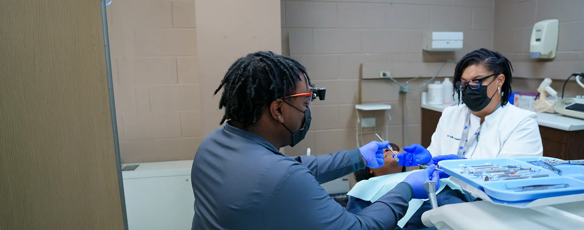 Student and instructor wearing gloves and masks perform a dental procedure on a patient in a training clinic.