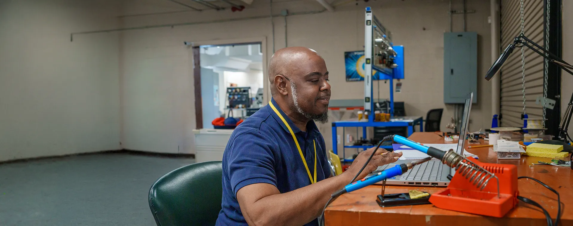 Instructor works at an electronics workstation with a laptop, soldering tools, and equipment in a training lab.