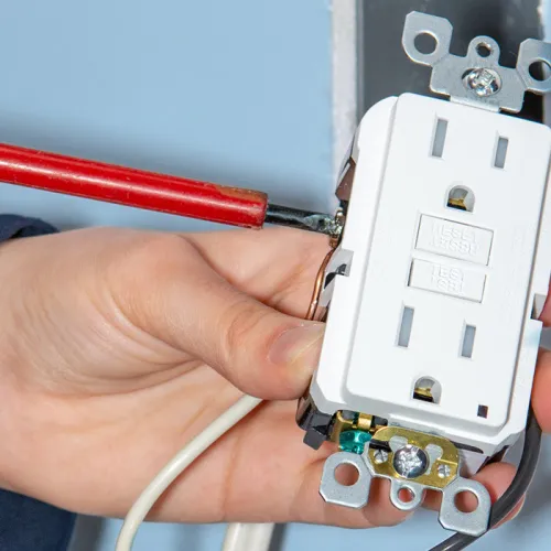 Hand uses a red screwdriver to secure wiring on a white GFCI electrical outlet mounted in a wall box