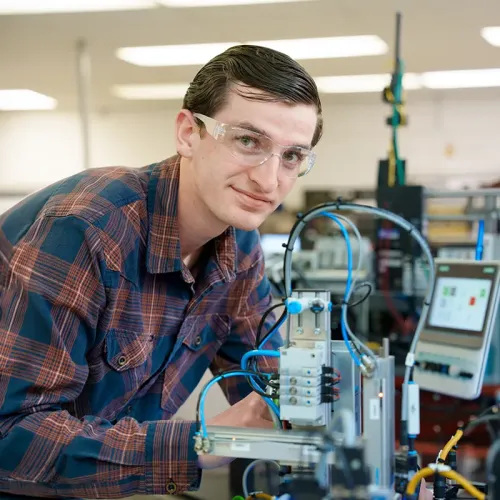 Instructor and student use a handheld controller beside a robotic training cell in an industrial automation lab.