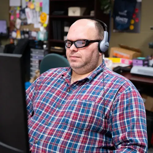Person wearing headphones and glasses working at computer, bulletin board and shelves with office items in background