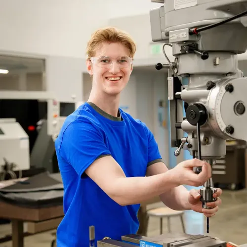Student wearing safety glasses operates a drill press in a machine tool training lab.