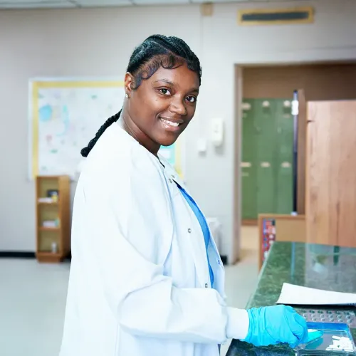Student in scrubs measures medication from a dispensing device with pharmacy shelves in the background.