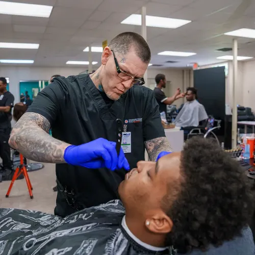 Student barber uses a straight razor during a shave practice session in a barbering training salon.