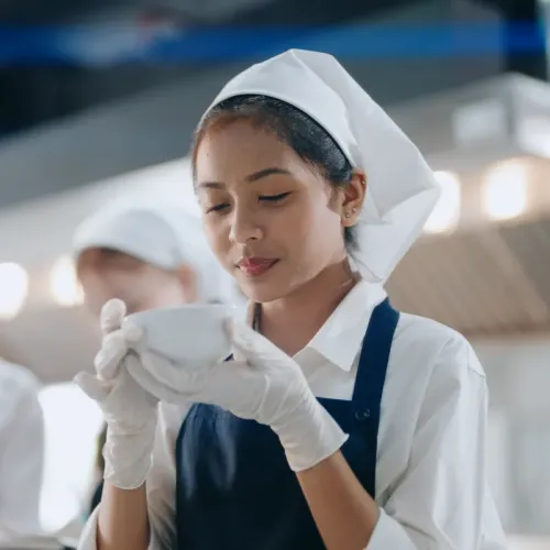 Student in a chef uniform and gloves holds a bowl while working in a commercial kitchen classroom.