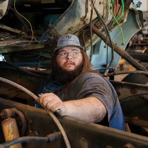 Student stands in a heavy equipment workshop holding a tool near a workbench and torch.