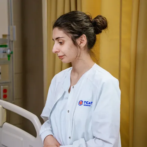 Student in a clinical uniform stands beside a hospital bed in a nursing training lab.