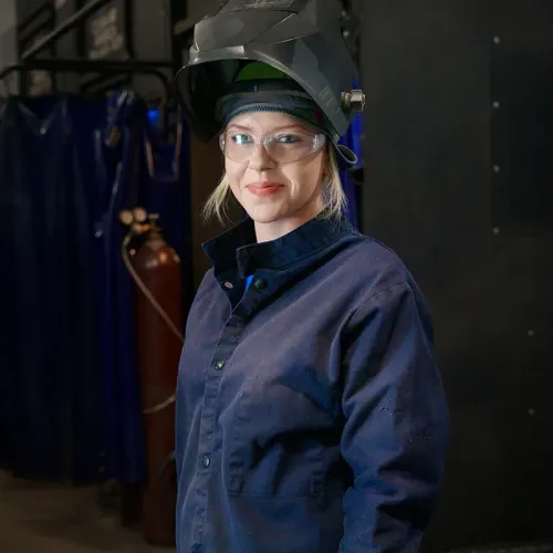 Student wearing safety glasses and a raised welding helmet stands in a welding training lab.