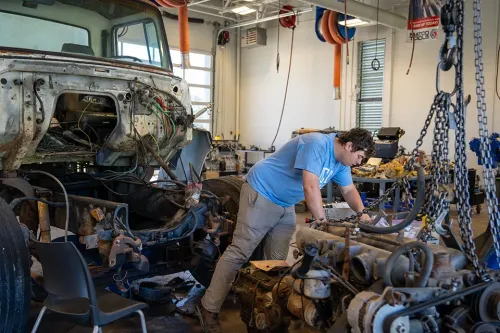 Student works beneath a truck cab, inspecting wiring and components on a vehicle frame.