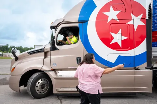 Instructor stands beside a CDL training truck and speaks with a student driver seated in the cab.