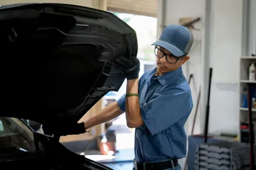 Student wearing gloves inspects and adjusts the underside of an open vehicle hood in a repair shop.
