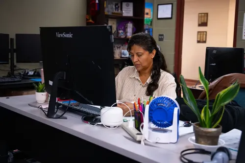 Person working at desk with desktop computer, small fan, plant, and office supplies, shelves and doorway in background