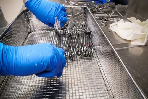 Gloved hands arrange surgical instruments in a wire sterilization tray on a stainless steel worktable.