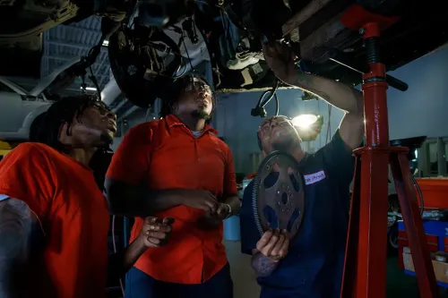 Instructor shows students vehicle components beneath a raised car in an automotive repair shop.