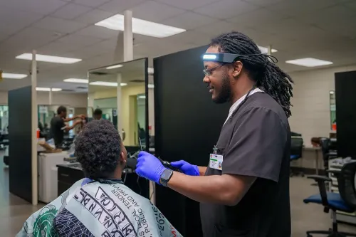 Student barber trims a client's hair with clippers in a barbering lab equipped with salon stations.