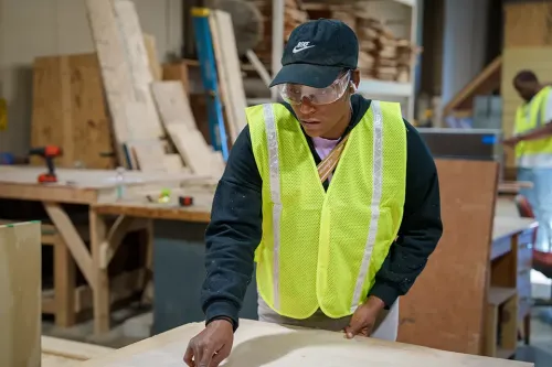 Student in safety glasses and reflective vest measures a wood panel in a construction training lab.