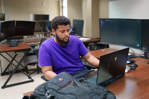 Student uses a laptop at a workstation surrounded by multiple computer monitors in a computer lab.
