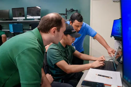 Student uses a laptop at a workstation surrounded by multiple computer monitors in a computer lab.