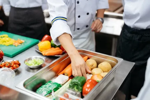Students in chef uniforms select vegetables and ingredients from a food preparation station in a training kitchen.