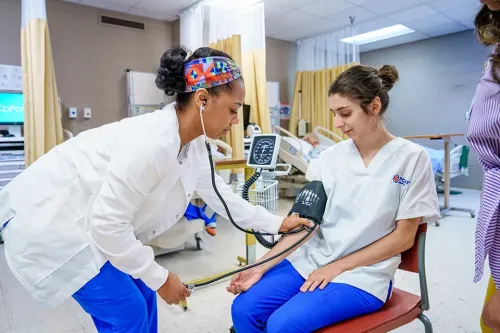 Student measures blood pressure on another student in a nursing skills lab with hospital equipment.