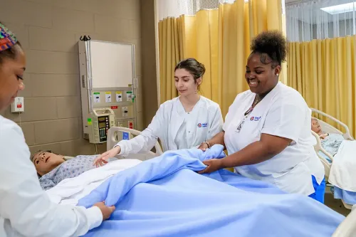 Nursing students make a hospital bed around a patient simulator in a clinical skills training lab.