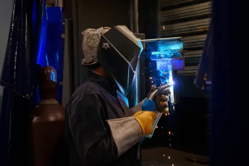 Student uses a welding torch on a metal workpiece while wearing a welding helmet and protective gloves.